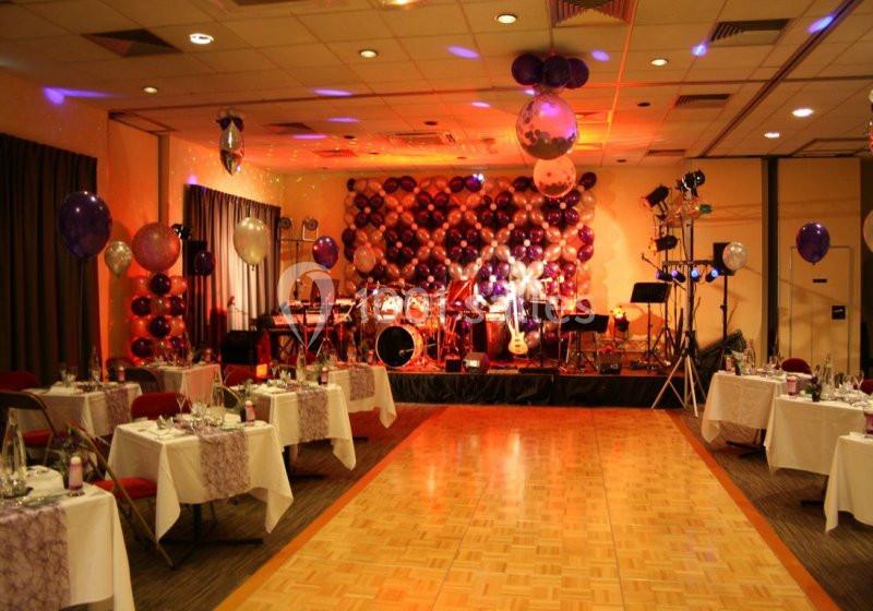 Salle décorée pour une fête avec ballons colorés, tables dressées et scène équipée pour un groupe musical.