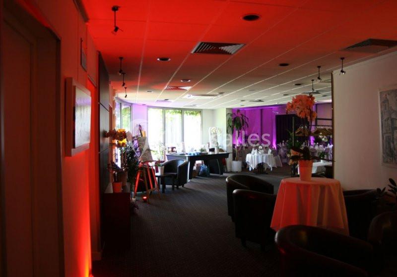 Salle lumineuse avec éclairage rouge et violet, tables dressées, fauteuils et décorations florales.