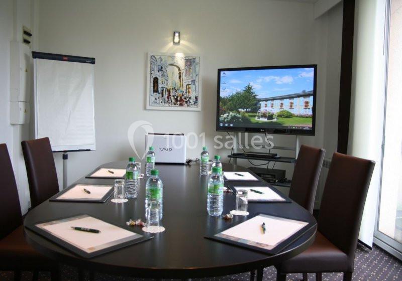 Salle de réunion avec table ovale, chaises, bloc-notes, bouteilles d'eau, écran allumé et paperboard.