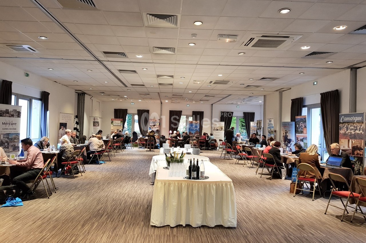 Salle de conférence avec des participants assis à des tables, discutant ou travaillant, entourés de stands promotionnels.