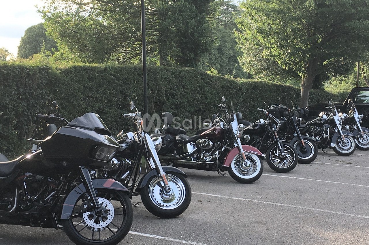 Plusieurs motos alignées sur un parking extérieur entouré de haies et d'arbres.