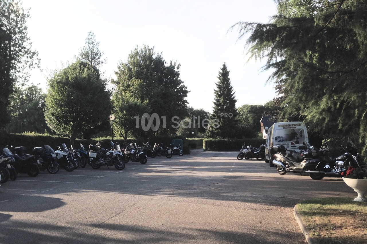 Plusieurs motos stationnées dans un parking entouré d'arbres, avec un véhicule utilitaire en arrière-plan.