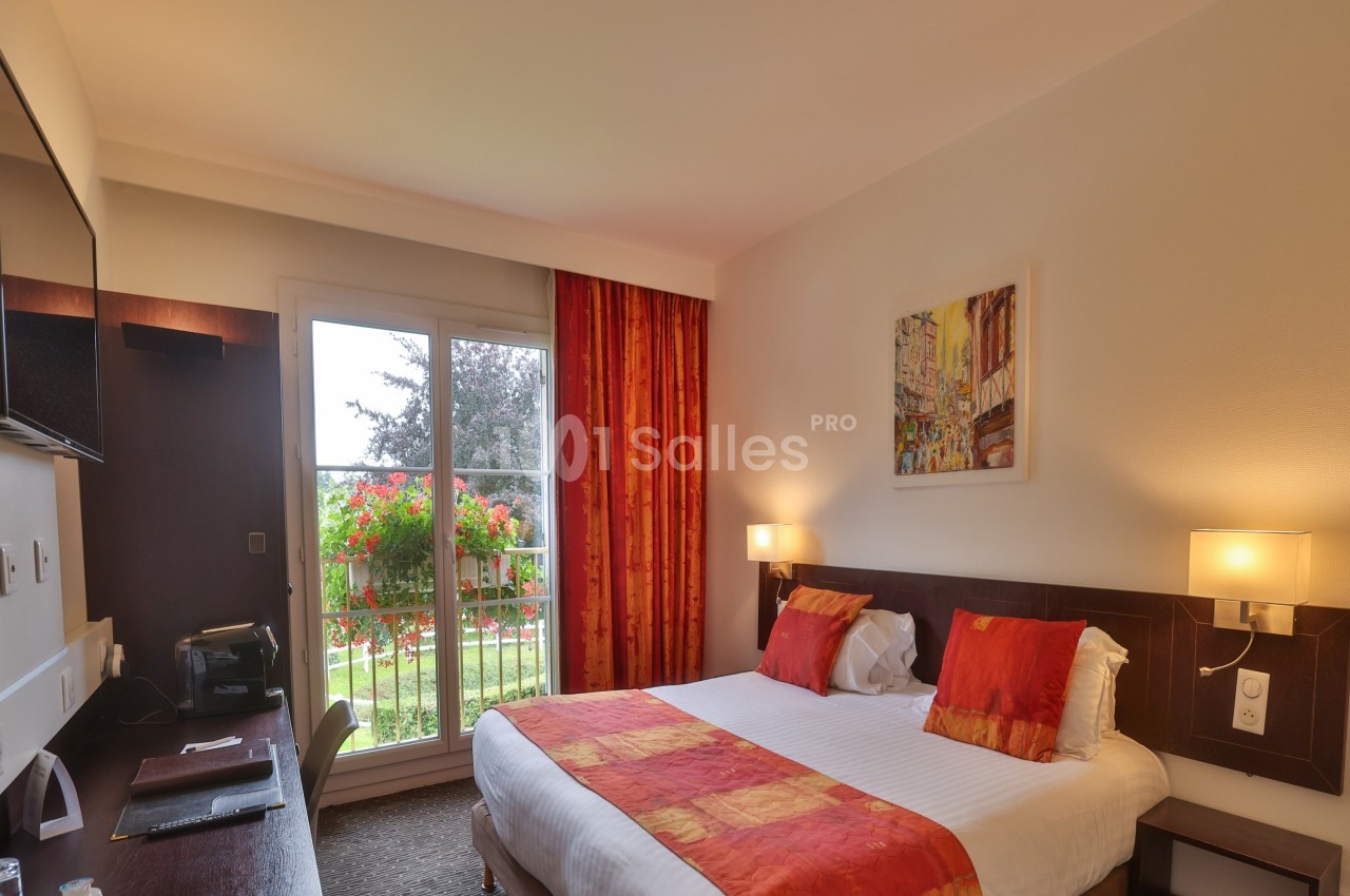Chambre d'hôtel lumineuse avec lit double, décorée de tons rouges et orange, donnant sur un balcon avec vue sur un jardin…
