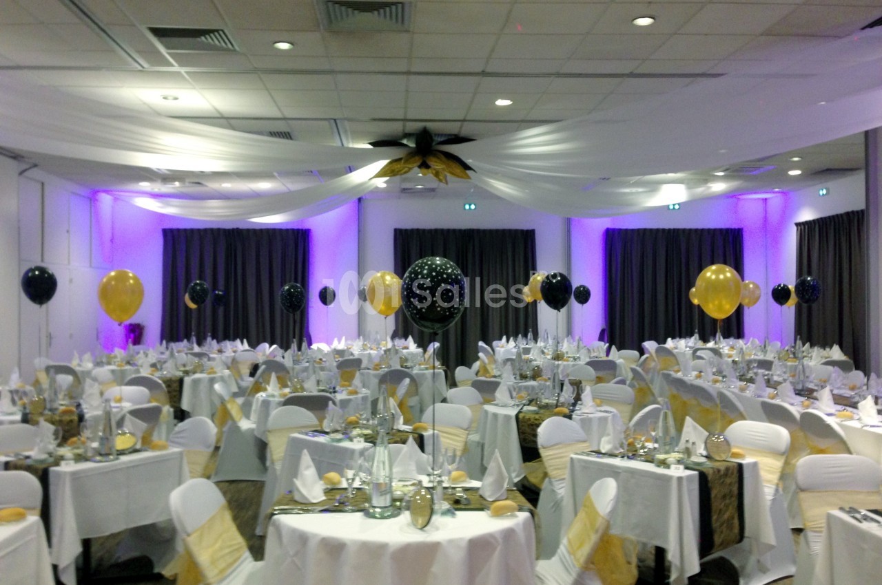Salle de réception décorée avec des ballons noirs et dorés, tables dressées avec nappes blanches et serviettes pliées.