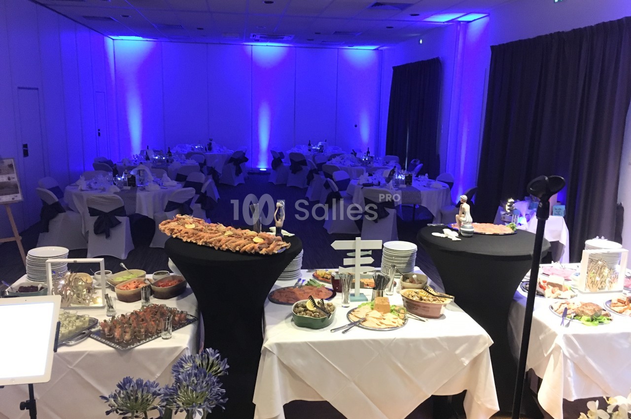 Salle de réception éclairée en bleu avec des tables dressées et un buffet varié au premier plan.
