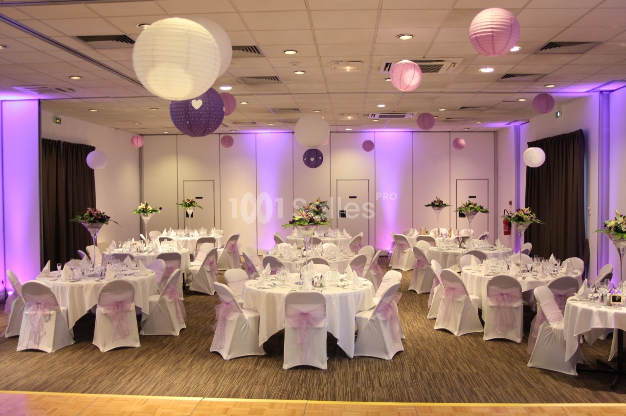 Salle de réception décorée avec des tables rondes, nappes blanches, chaises ornées de rubans roses et lanternes suspendues.