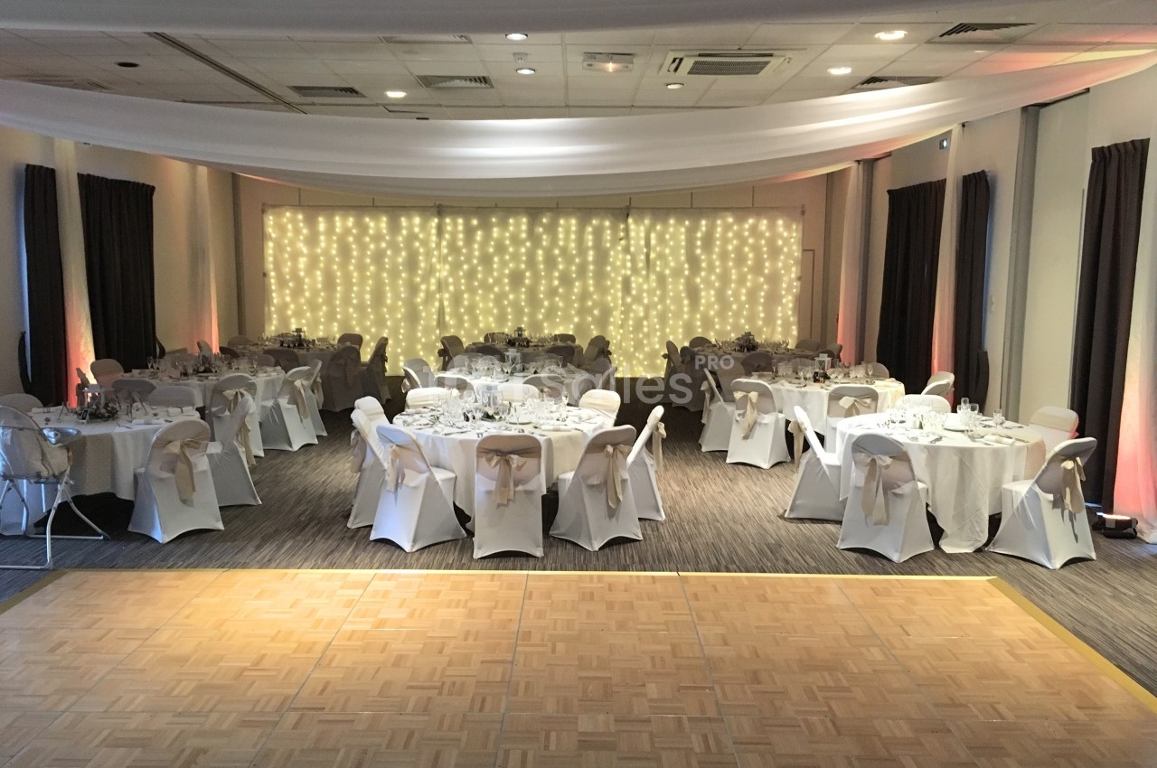 Salle de réception décorée avec des tables rondes dressées et un mur illuminé de guirlandes lumineuses.