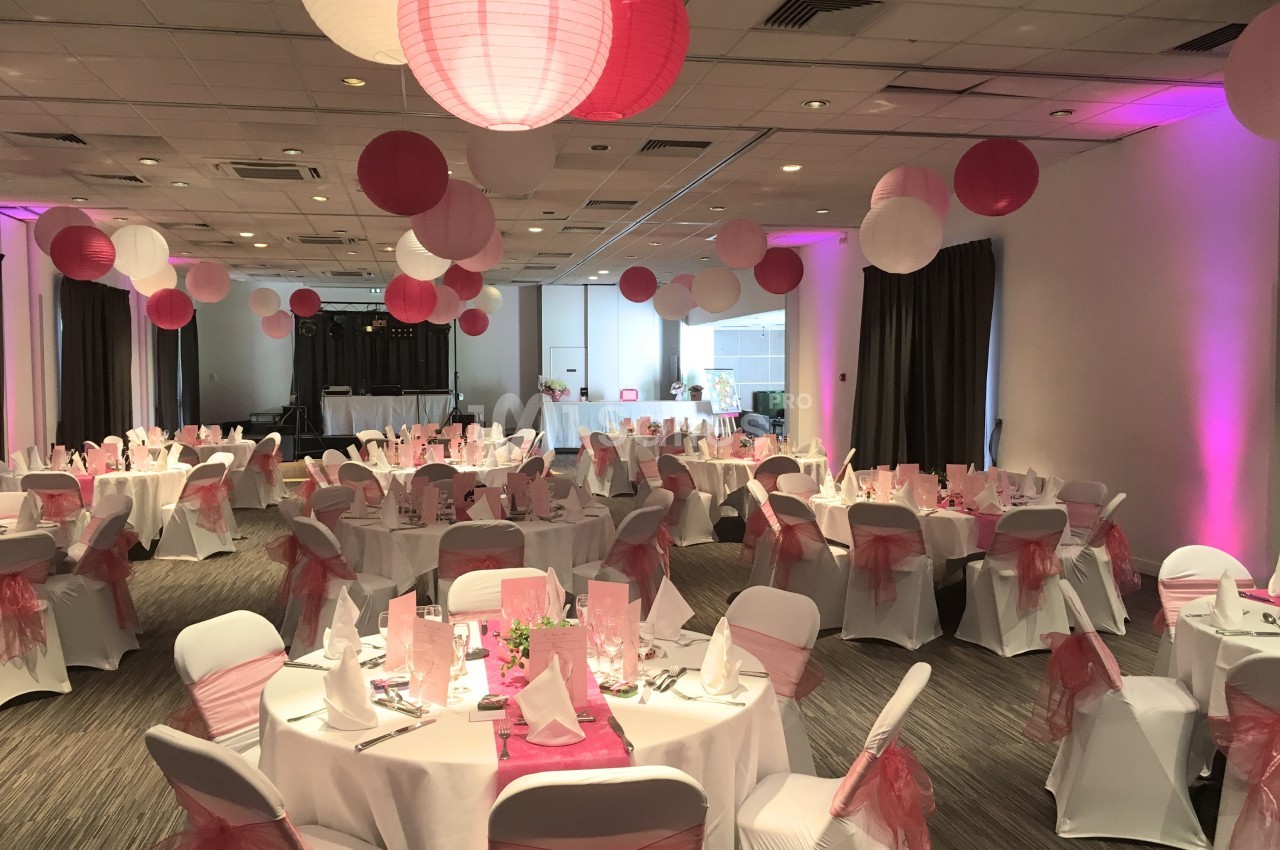 Salle de réception décorée avec des nappes blanches, des rubans roses et des lanternes suspendues roses et blanches.