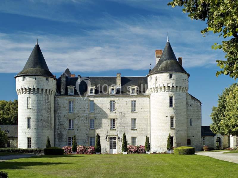 Château en pierre blanche avec deux tours rondes, entouré de verdure et d'arbustes fleuris sous un ciel bleu.