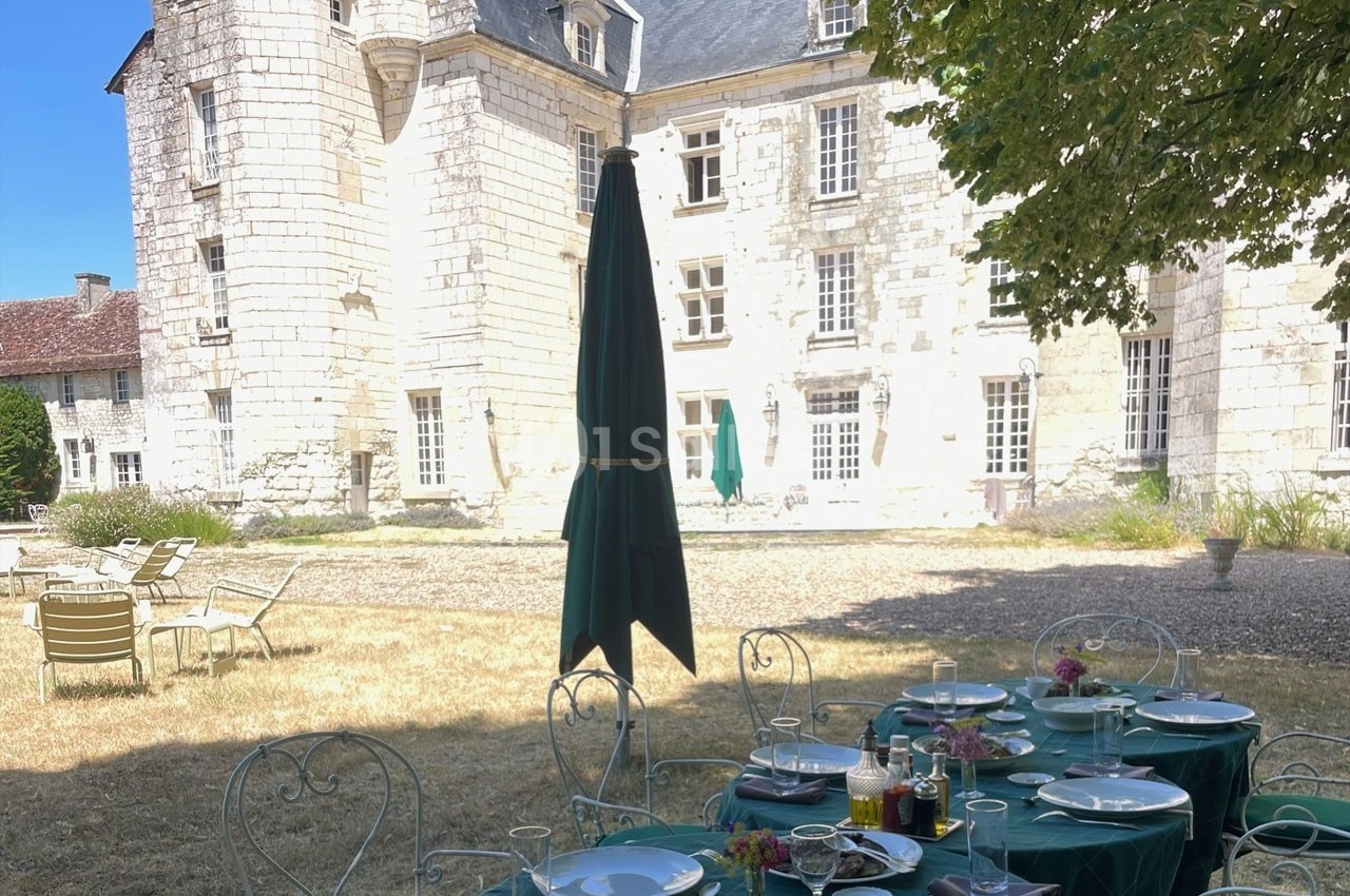 Table dressée avec vaisselle et fleurs sous un arbre, devant un château en pierre blanche par une journée ensoleillée.