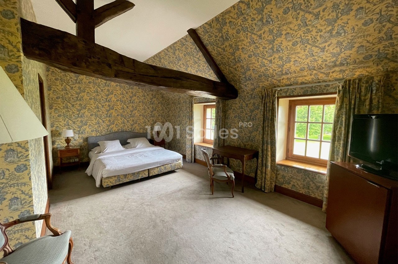 Chambre spacieuse avec lit double, poutres apparentes, murs tapissés, fenêtres donnant sur un jardin verdoyant.