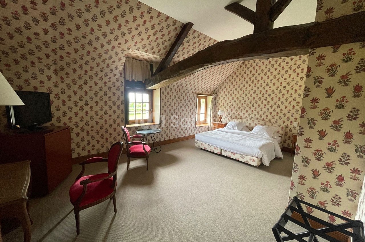 Chambre mansardée avec poutres apparentes, lit double, mobilier classique et murs tapissés de motifs floraux.
