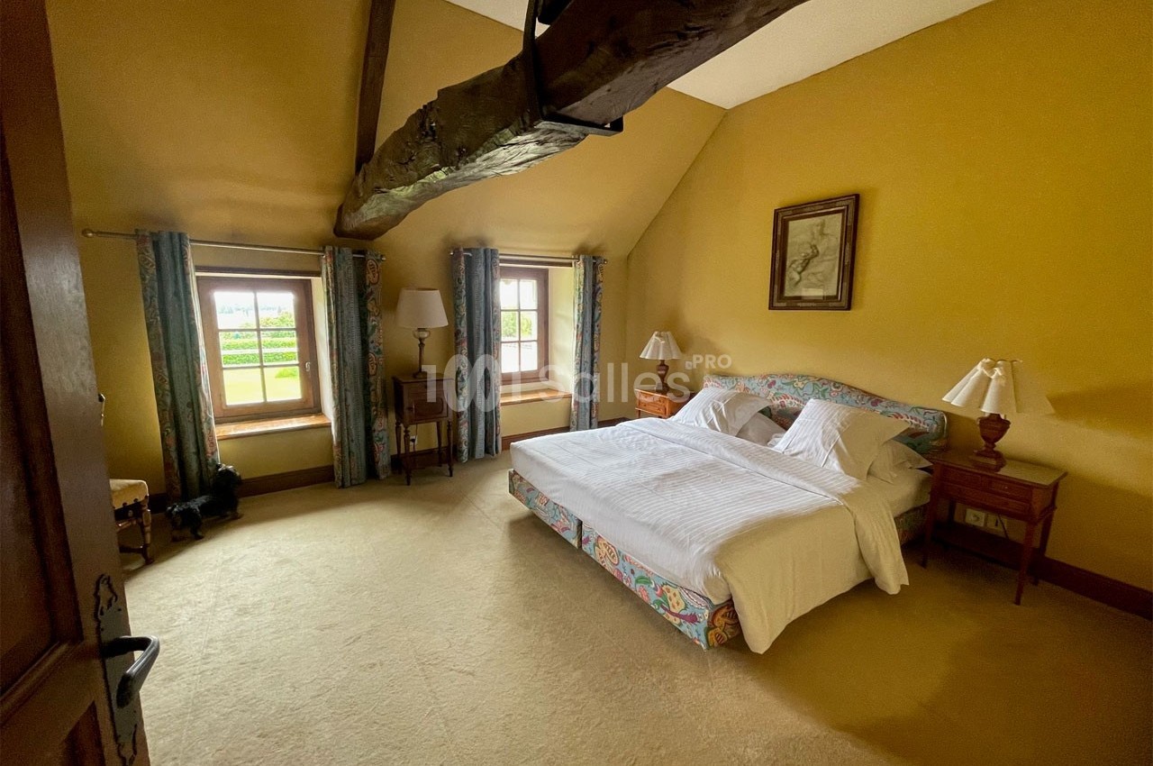 Chambre avec lit double, poutre apparente, murs jaunes, deux fenêtres avec rideaux et mobilier en bois.
