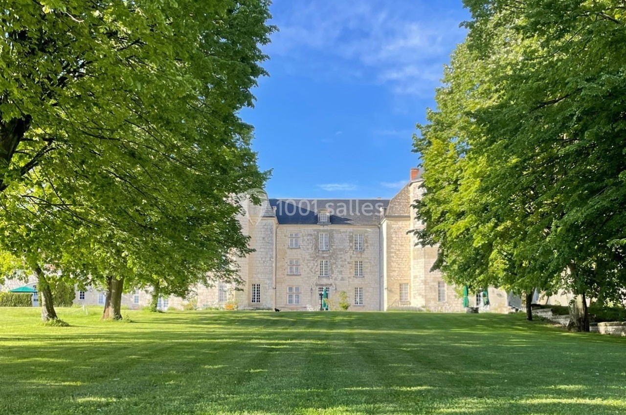 Façade d'un château en pierre entouré d'arbres et d'une pelouse verdoyante sous un ciel bleu.