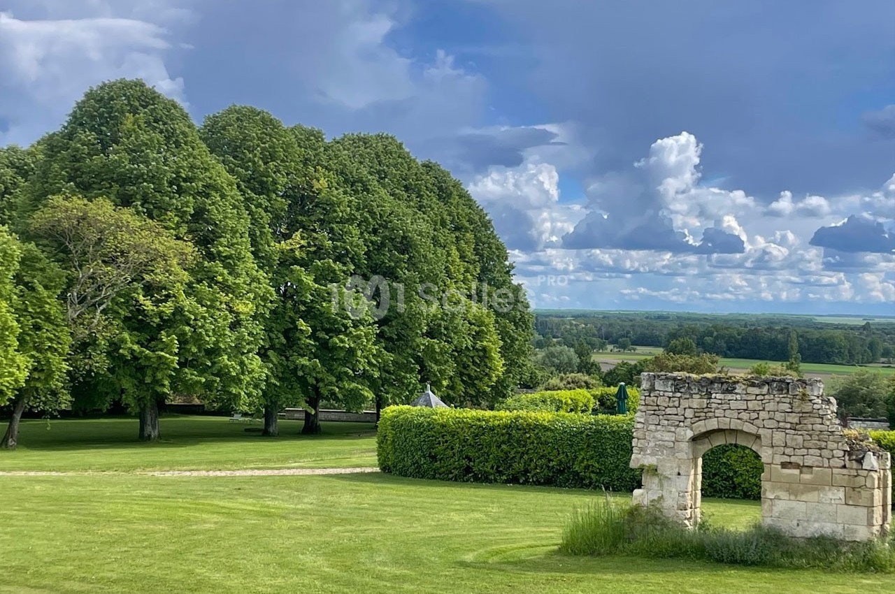 Ruine en pierre dans un parc verdoyant avec pelouse, arbres imposants et ciel partiellement nuageux en arrière-plan.
