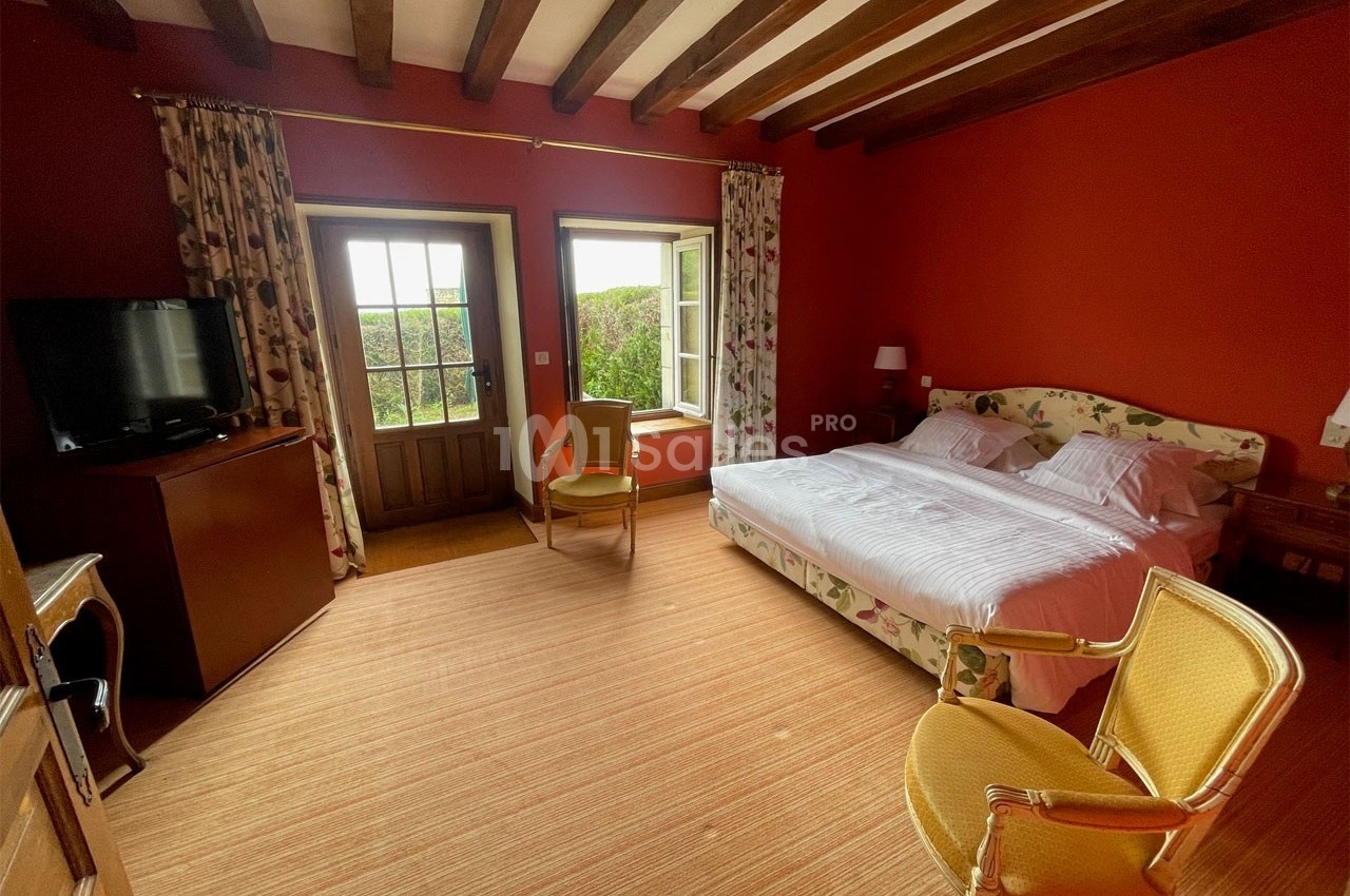 Chambre avec murs rouges, lit double, fauteuils, télévision, fenêtres donnant sur un jardin et poutres apparentes au plafond.