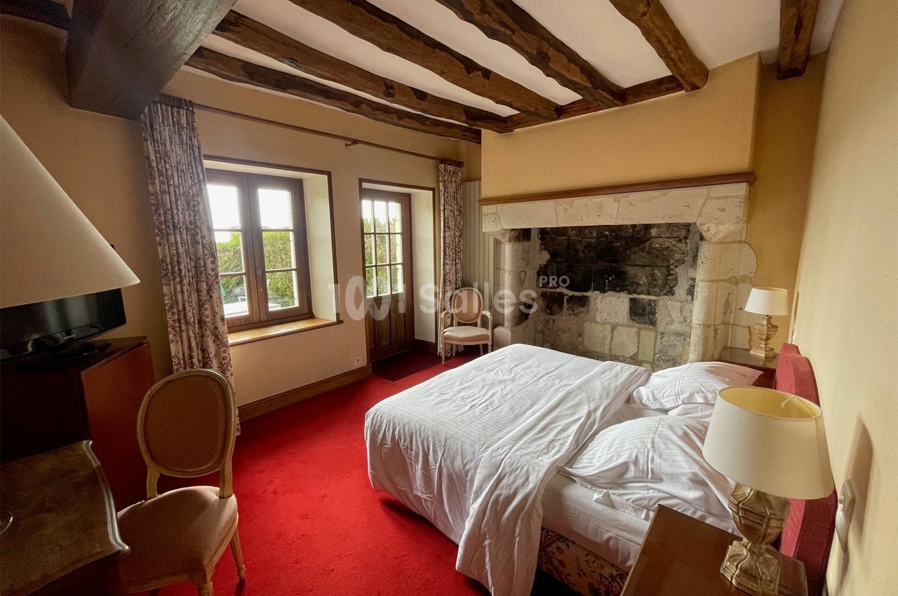 Chambre avec lit double, poutres apparentes, cheminée en pierre, moquette rouge et fenêtres donnant sur un jardin.