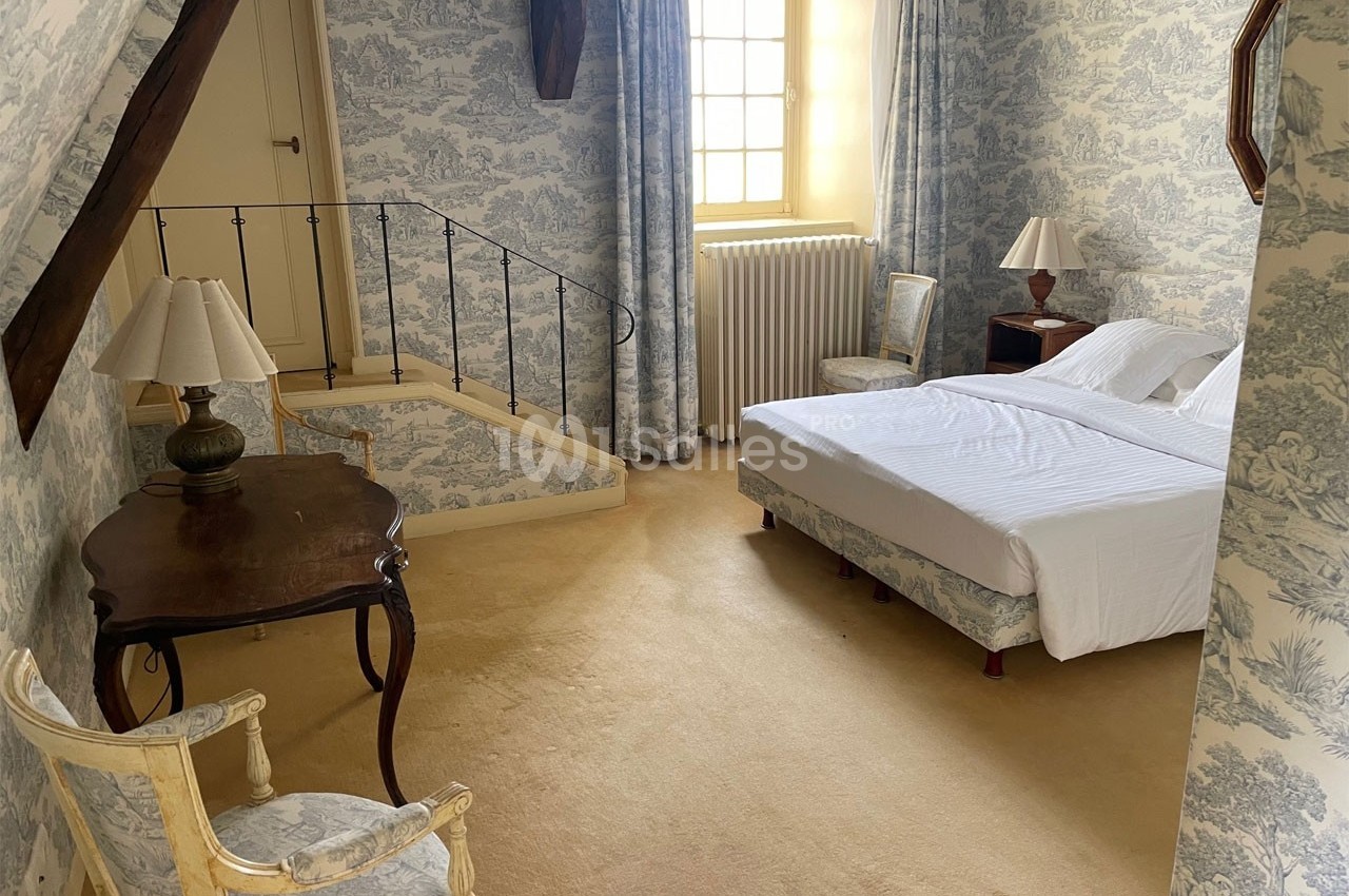 Chambre lumineuse avec lit double, mobilier classique, murs et rideaux à motifs, et escalier menant à une porte.