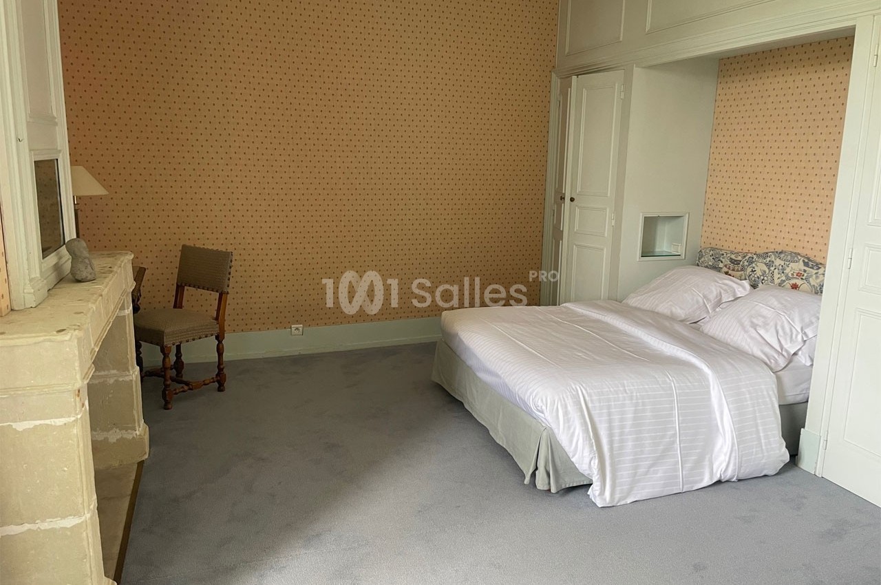 Chambre avec un lit double, draps blancs, cheminée en pierre, chaise en bois et murs tapissés de motifs discrets.