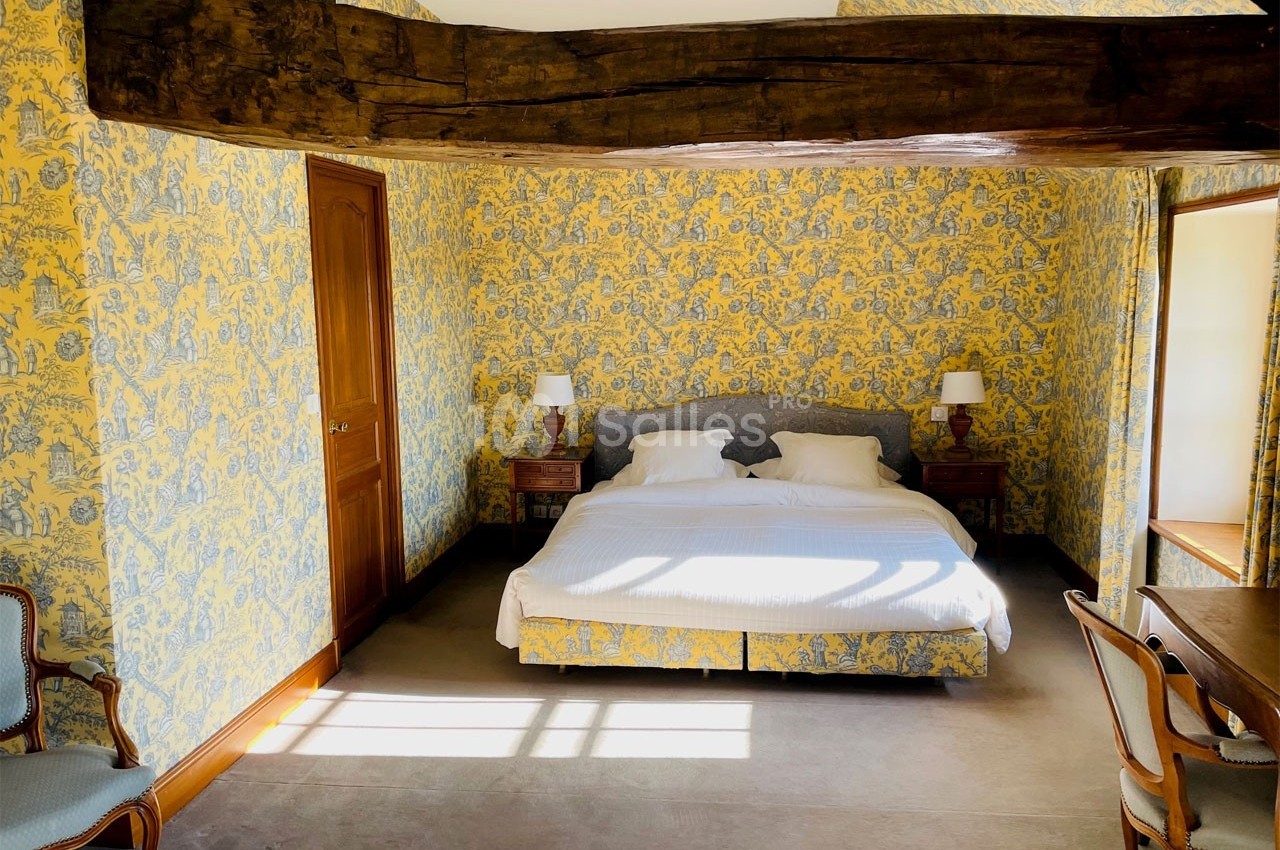 Chambre lumineuse avec lit double, poutre apparente, murs tapissés de motifs jaunes et bleus, et mobilier classique.