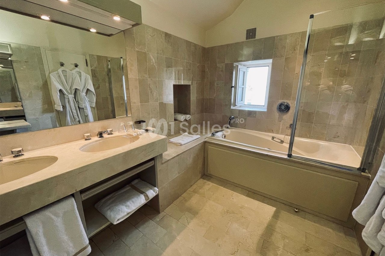 Salle de bain avec double vasque, baignoire encastrée, murs en carrelage beige et peignoirs accrochés.