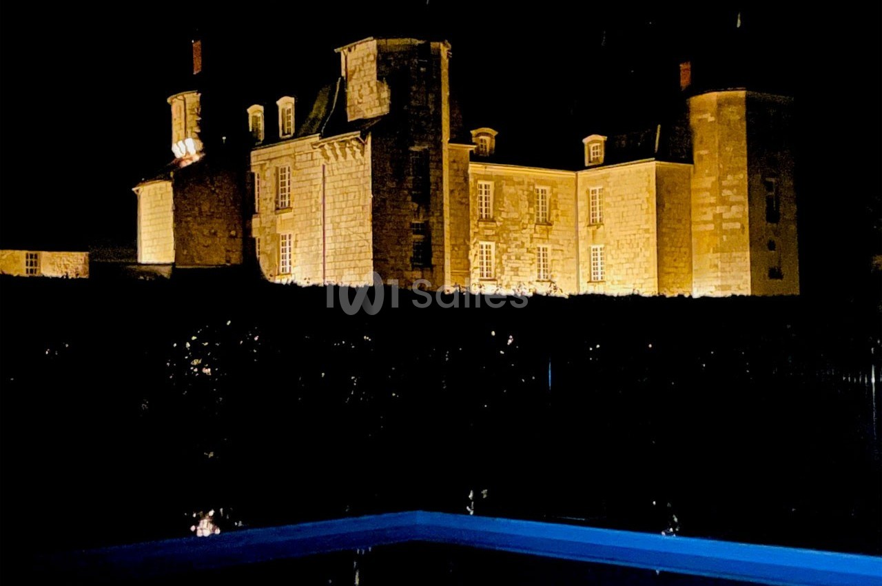 Château en pierre éclairé la nuit, avec une piscine illuminée au premier plan.