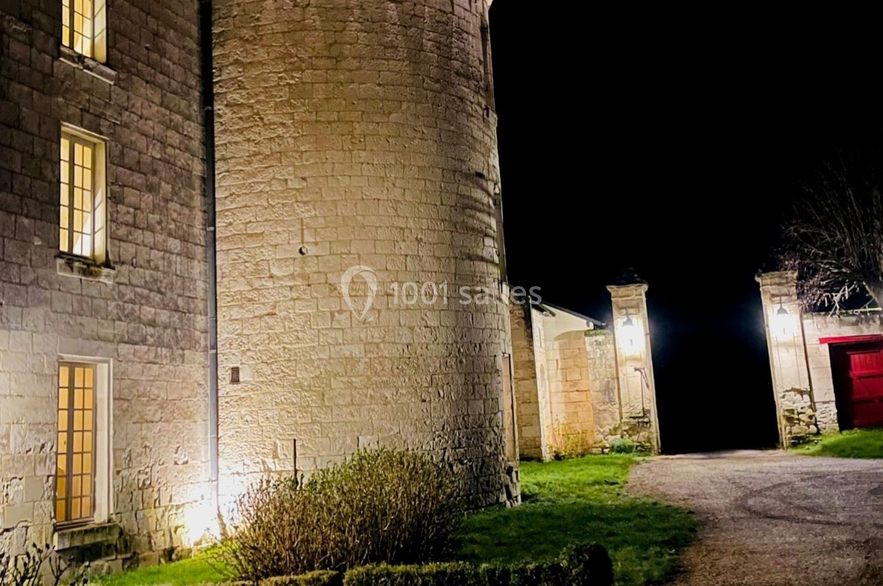 Façade d'un bâtiment en pierre éclairé la nuit, avec une tour ronde et une entrée ouverte sur un chemin.