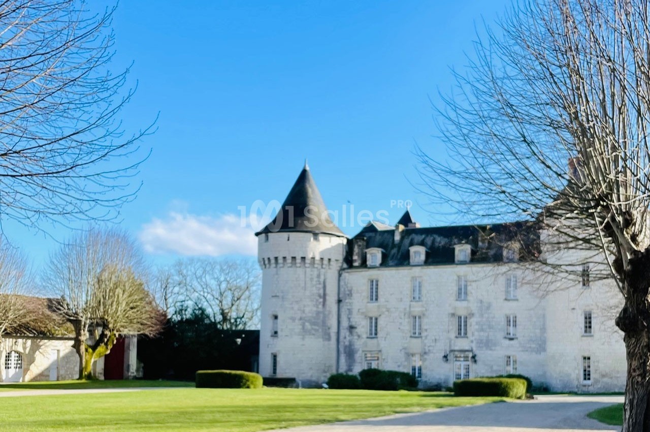 Château en pierre blanche entouré d'arbres dénudés, avec pelouse et ciel bleu en arrière-plan.