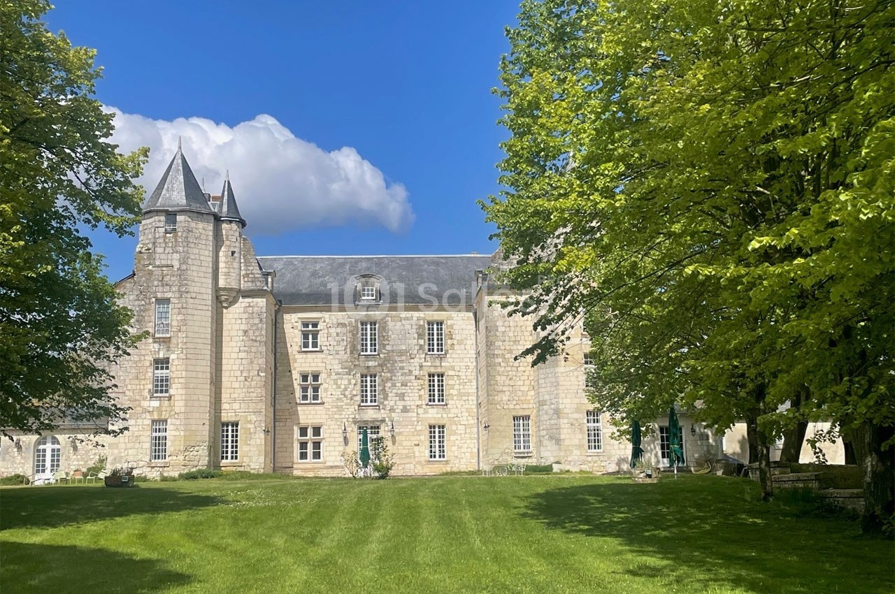 Façade d'un château en pierre entouré d'arbres, avec une pelouse verdoyante au premier plan sous un ciel bleu.