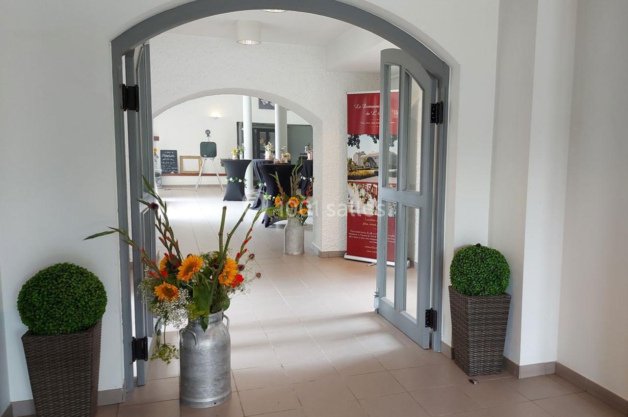 Entrée lumineuse avec porte ouverte, décorée de plantes et fleurs, menant à une salle aménagée pour un événement.