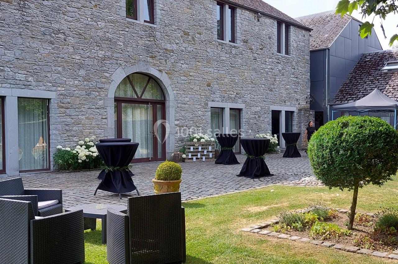 Cour pavée avec façade en pierre, tables hautes noires, mobilier de jardin et pelouse aménagée.