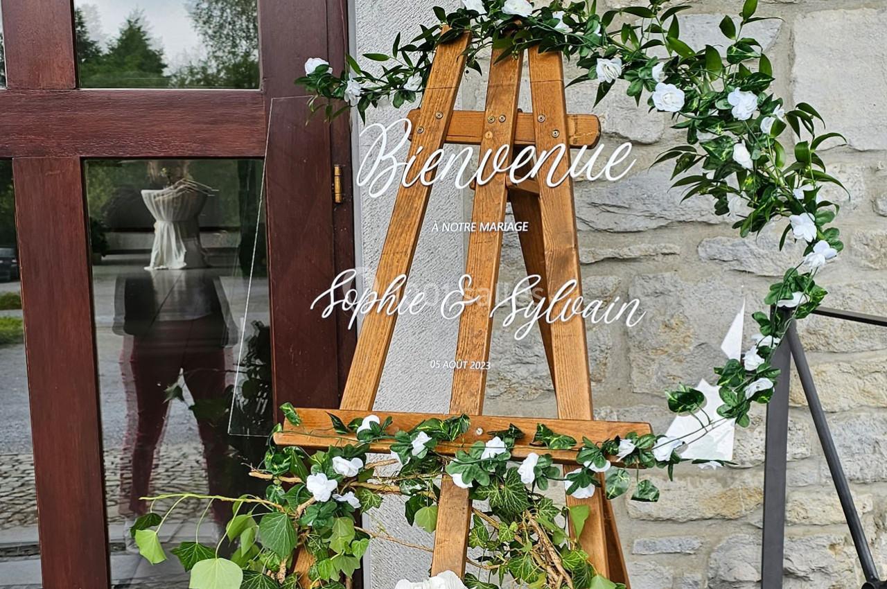 Chevalet en bois décoré de feuillage et fleurs blanches, avec un panneau ’Bienvenue à notre mariage’ devant un mur en pierre.