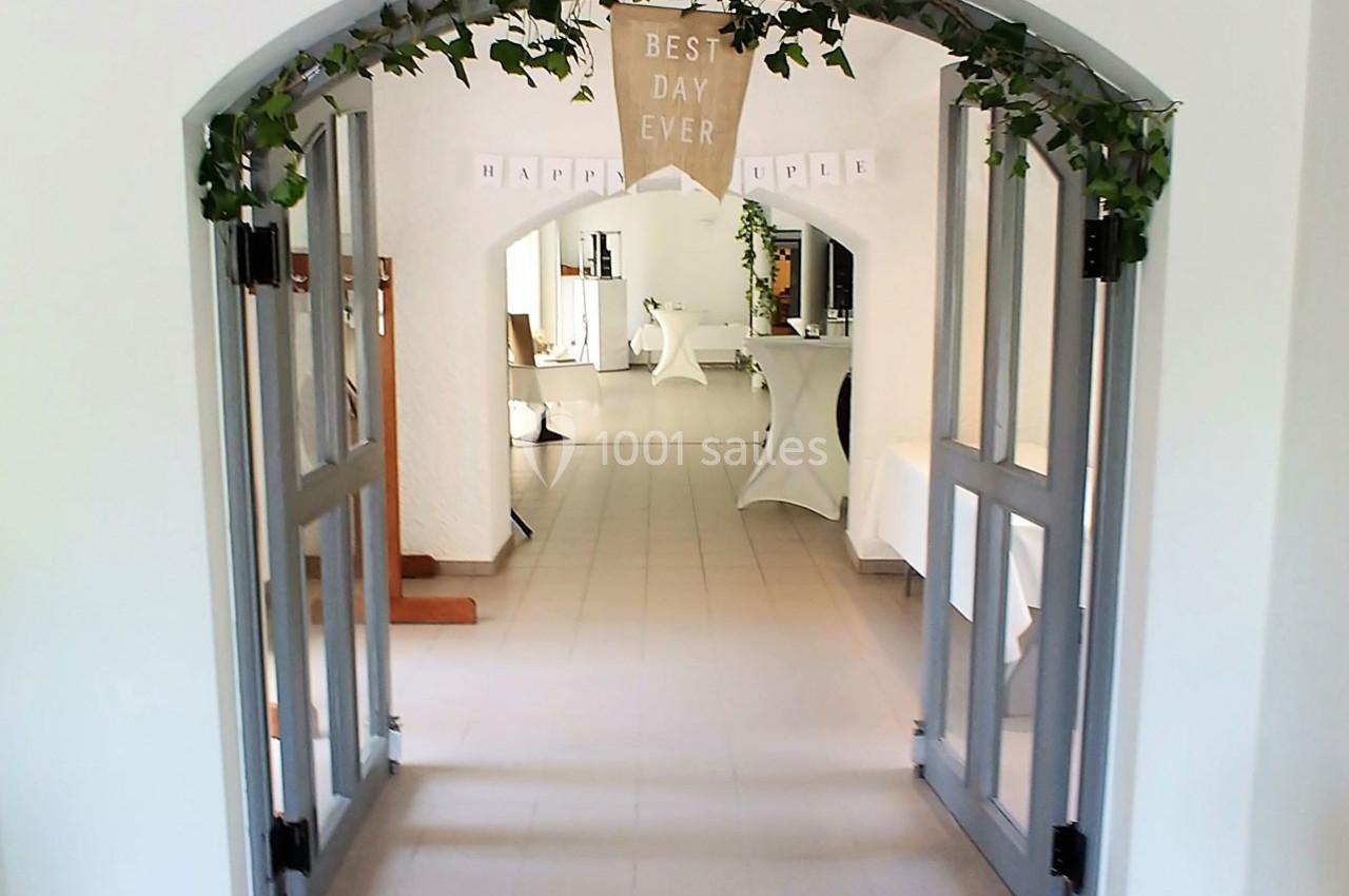 Entrée d'une salle décorée pour un événement, avec une arche ornée de feuillage et une bannière ’Best Day Ever’.