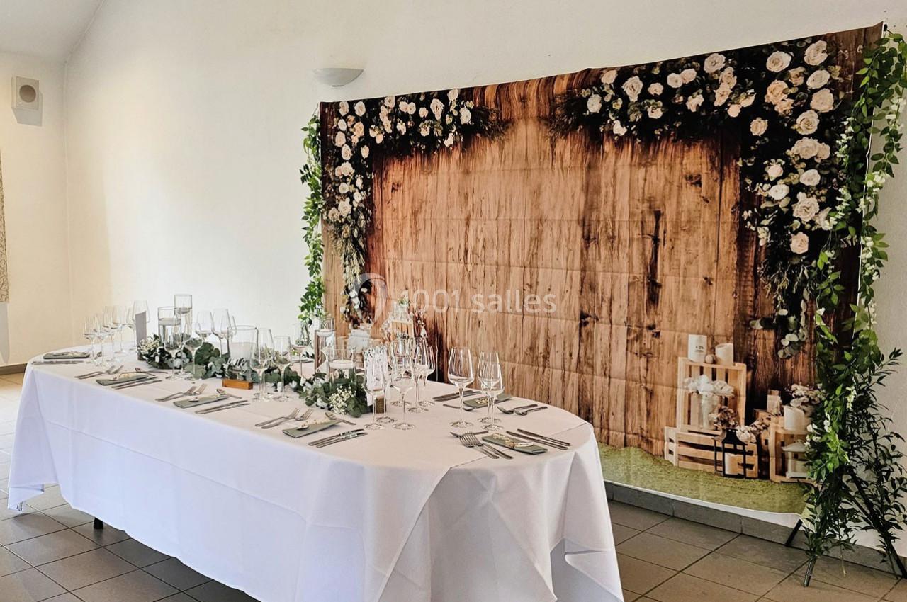 Table décorée pour un événement avec nappes blanches, verrerie élégante et fond en bois orné de fleurs blanches.