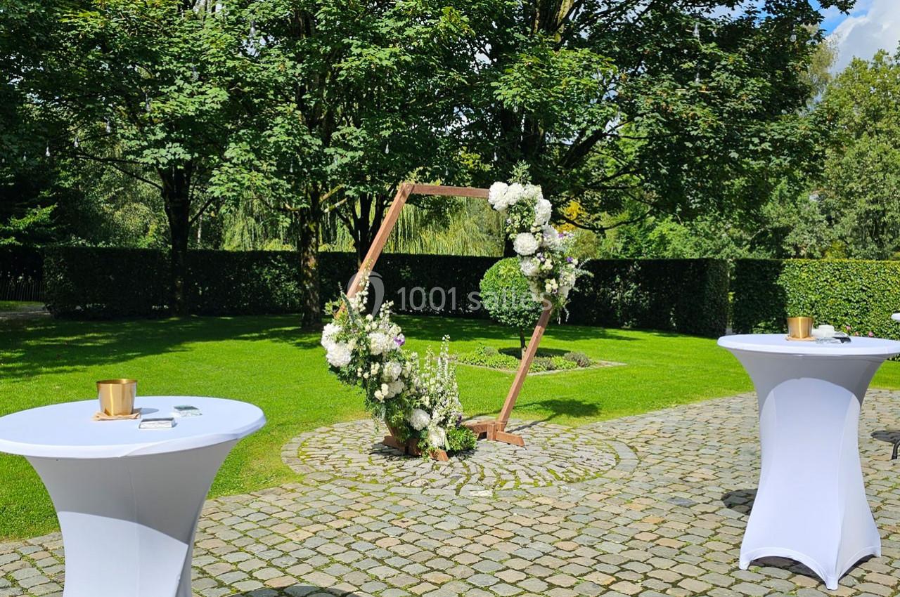 Arche hexagonale décorée de fleurs blanches dans un jardin, entourée de tables hautes drapées de blanc.