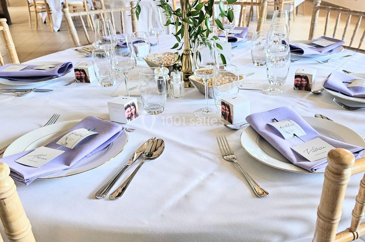 Table dressée avec une nappe blanche, serviettes lavande, couverts, verres, et décorations végétales au centre.