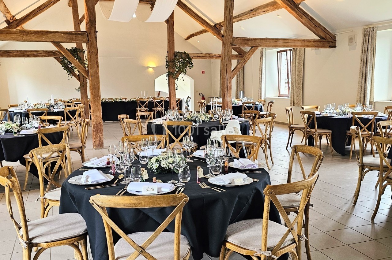 Salle de réception lumineuse avec tables rondes dressées, nappes noires et chaises en bois, décorée pour un événement.