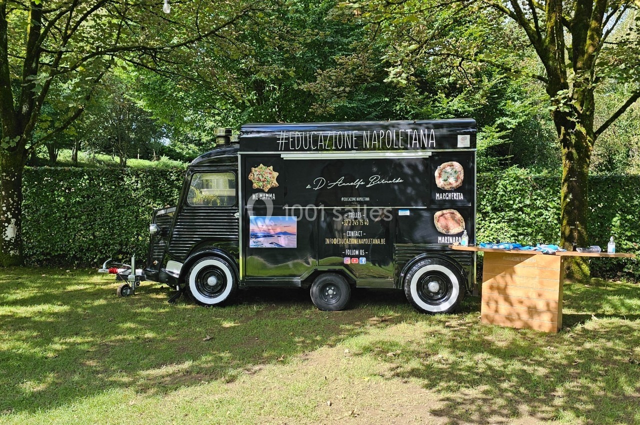 Camion noir de type food truck stationné sur une pelouse, entouré d'arbres, avec une table en bois à côté.