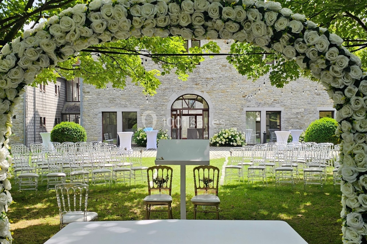 Arche de roses blanches devant des chaises disposées pour une cérémonie en plein air, avec un bâtiment en pierre en arrière…
