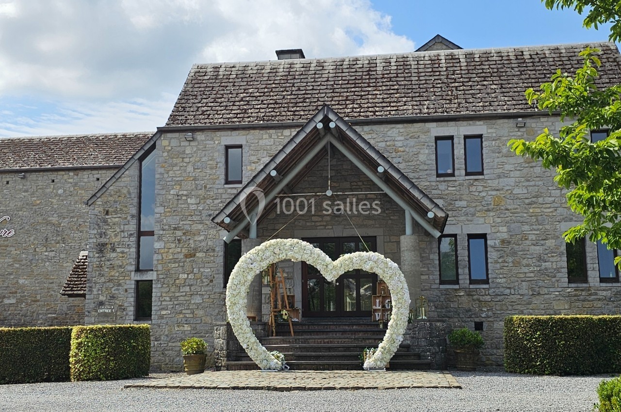 Façade en pierre d'un bâtiment avec une arche en forme de cœur décorée de fleurs blanches devant l'entrée.