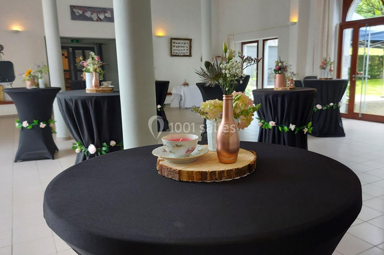 Décoration de table avec une bouteille dorée, des fleurs et une tasse sur une rondelle de bois dans une salle de réception.