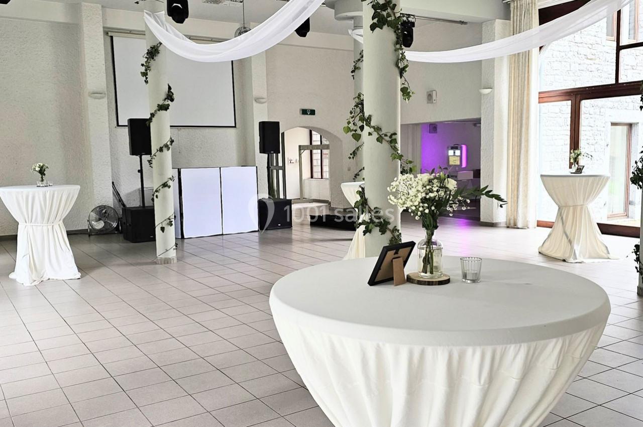 Salle de réception lumineuse décorée de drapés blancs et de verdure, avec des tables hautes recouvertes de nappes.