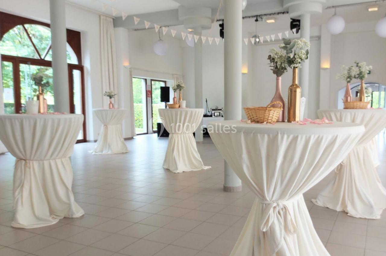 Salle lumineuse décorée pour un événement, avec des tables hautes drapées de blanc et des vases fleuris.