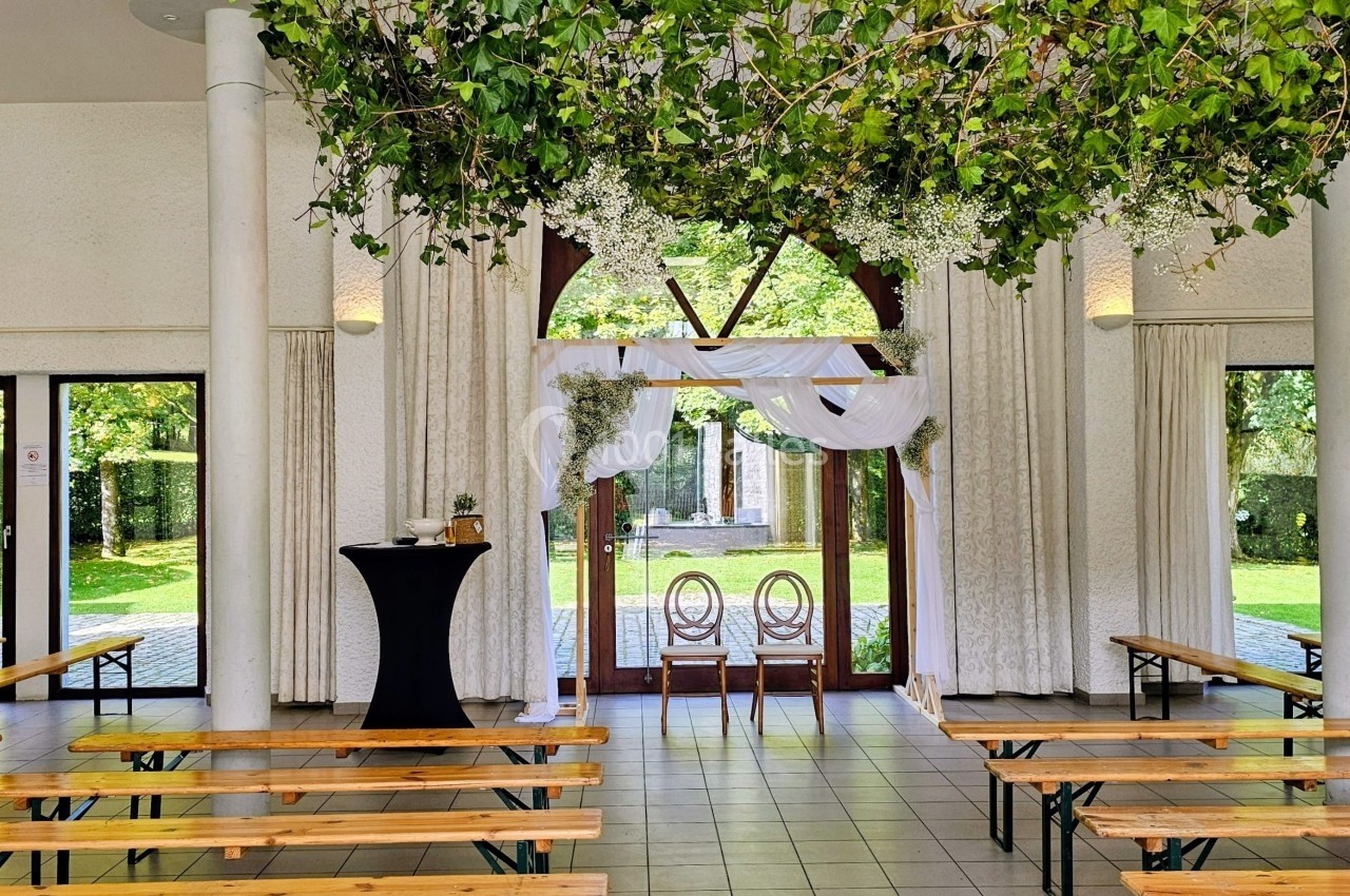 Salle de réception lumineuse avec bancs en bois, décor floral suspendu et vue sur un jardin à travers de grandes baies…