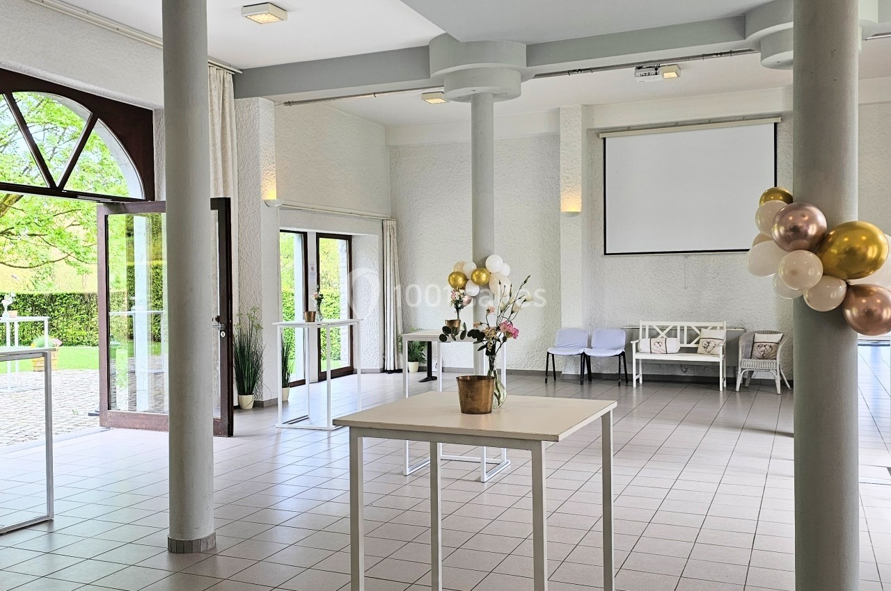 Salle lumineuse avec grandes fenêtres, colonnes blanches, décorations de ballons et mobilier minimaliste.