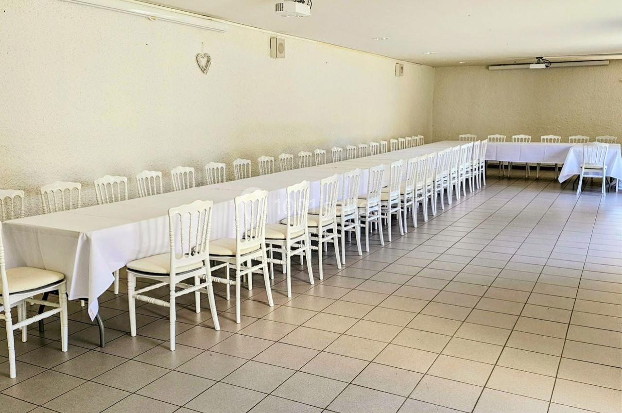 Salle lumineuse avec deux longues tables blanches alignées, entourées de chaises blanches, sur un sol carrelé.
