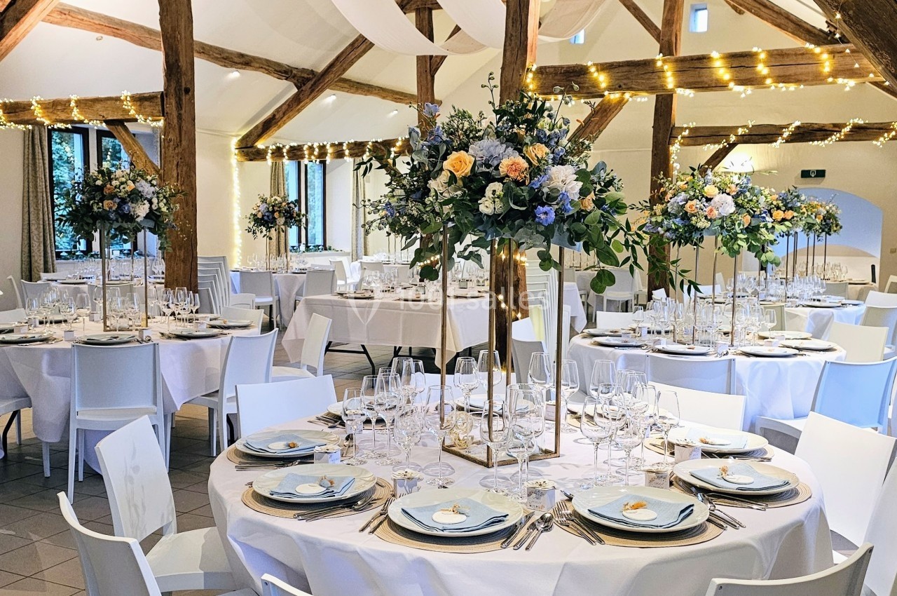 Salle de réception décorée avec des tables rondes, nappes blanches, vaisselle élégante et centres de table fleuris.