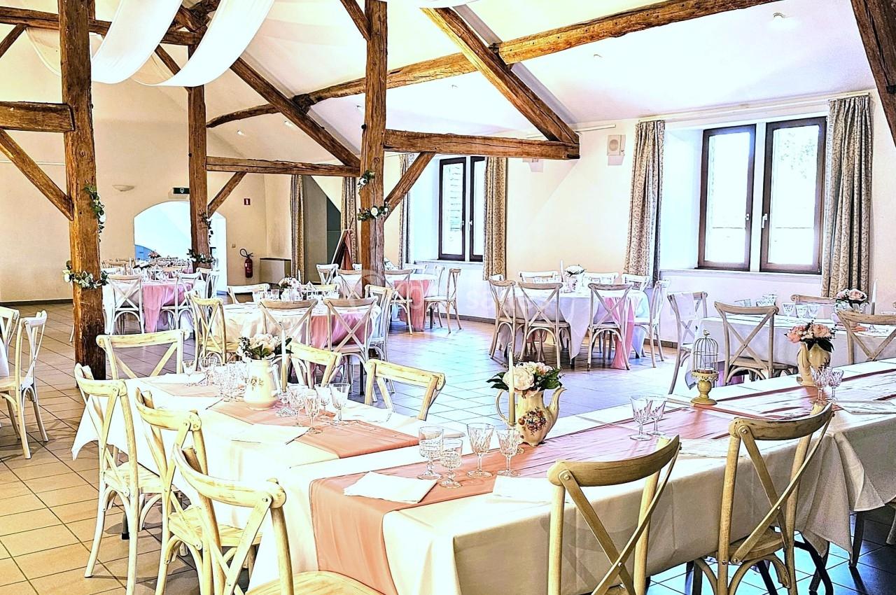 Salle de réception lumineuse avec poutres apparentes, tables décorées de nappes blanches et chemins de table roses.