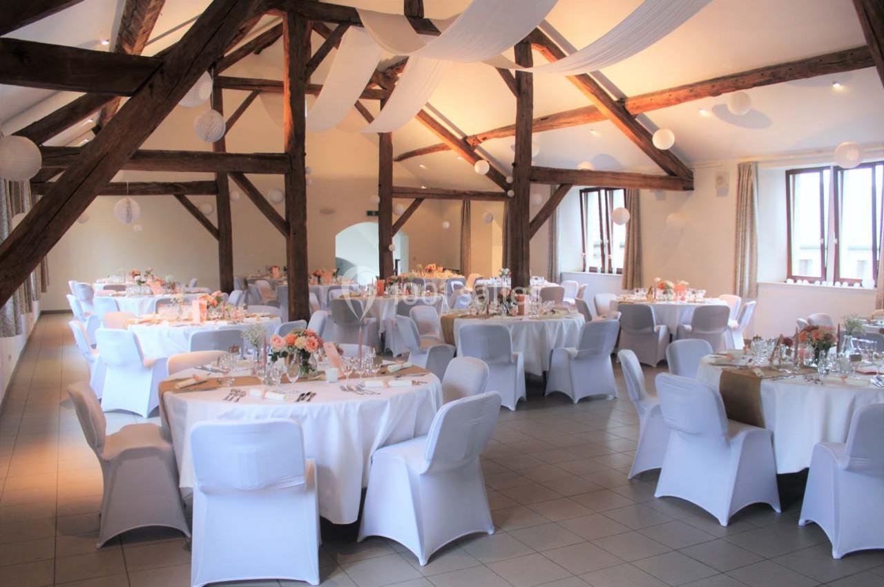 Salle de réception lumineuse avec des tables rondes décorées, des chaises blanches et des poutres apparentes au plafond.
