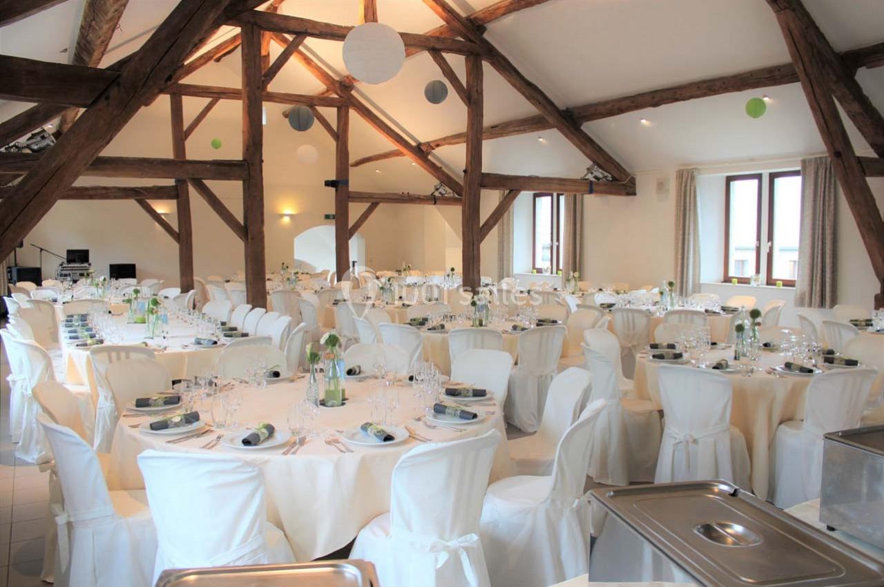 Salle de réception lumineuse avec des tables rondes dressées, nappes blanches et poutres apparentes au plafond.